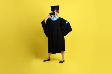 Mavi mezuniyet elbisesi ve şapkalı, sarı arka planda izole cep telefonu bulunduran bir kız öğrenci. Gen Z, yeni mezun, eğitim, ilk iş