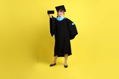 Mavi mezuniyet elbisesi ve şapkalı, sarı arka planda izole cep telefonu bulunduran bir kız öğrenci. Gen Z, yeni mezun, eğitim, ilk iş
