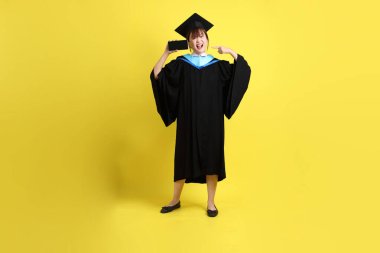 Mavi mezuniyet elbisesi ve şapkalı, sarı arka planda izole cep telefonu bulunduran bir kız öğrenci. Gen Z, yeni mezun, eğitim, ilk iş