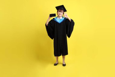 Mavi mezuniyet elbisesi ve şapkalı, sarı arka planda izole cep telefonu bulunduran bir kız öğrenci. Gen Z, yeni mezun, eğitim, ilk iş