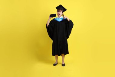 Mavi mezuniyet elbisesi ve şapkalı, sarı arka planda izole cep telefonu bulunduran bir kız öğrenci. Gen Z, yeni mezun, eğitim, ilk iş
