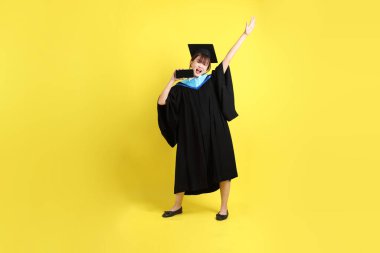 Mavi mezuniyet elbisesi ve şapkalı, sarı arka planda izole cep telefonu bulunduran bir kız öğrenci. Gen Z, yeni mezun, eğitim, ilk iş