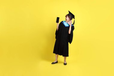 Sarı arka planda izole edilmiş selfie çekme jesti ile mavi mezuniyet elbisesi ve şapkalı bir üniversite öğrencisi. Gen Z, yeni mezun, eğitim, ilk iş