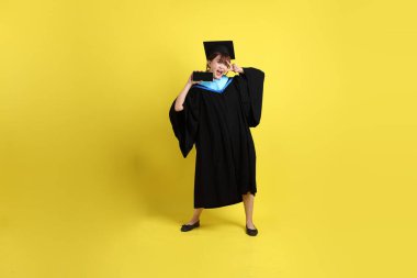 Mavi mezuniyet elbisesi ve şapkalı, sarı arka planda izole cep telefonu bulunduran bir kız öğrenci. Gen Z, yeni mezun, eğitim, ilk iş