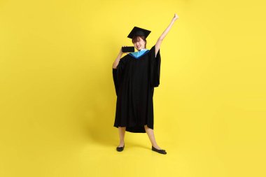 Mavi mezuniyet elbisesi ve şapkalı, sarı arka planda izole cep telefonu bulunduran bir kız öğrenci. Gen Z, yeni mezun, eğitim, ilk iş