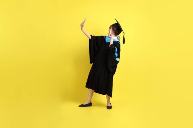 Sarı arka planda izole edilmiş selfie çekme jesti ile mavi mezuniyet elbisesi ve şapkalı bir üniversite öğrencisi. Gen Z, yeni mezun, eğitim, ilk iş