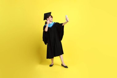 Sarı arka planda izole edilmiş selfie çekme jesti ile mavi mezuniyet elbisesi ve şapkalı bir üniversite öğrencisi. Gen Z, yeni mezun, eğitim, ilk iş