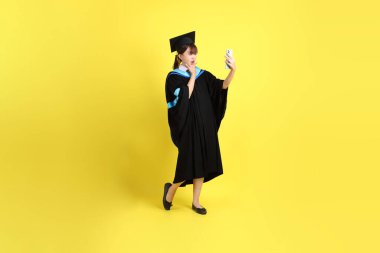 Sarı arka planda izole edilmiş selfie çekme jesti ile mavi mezuniyet elbisesi ve şapkalı bir üniversite öğrencisi. Gen Z, yeni mezun, eğitim, ilk iş