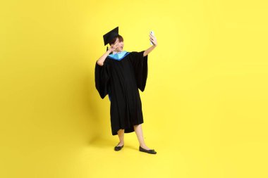 Sarı arka planda izole edilmiş selfie çekme jesti ile mavi mezuniyet elbisesi ve şapkalı bir üniversite öğrencisi. Gen Z, yeni mezun, eğitim, ilk iş