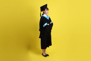 Mavi mezuniyet elbisesi ve şapkalı, ayakta durma ve kollarını çaprazlama hareketleriyle sarı arka planda izole edilmiş bir üniversite öğrencisi. Gen Z, yeni mezun, eğitim, ilk iş