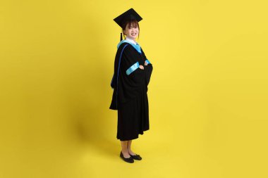 Mavi mezuniyet elbisesi ve şapkalı, ayakta durma ve kollarını çaprazlama hareketleriyle sarı arka planda izole edilmiş bir üniversite öğrencisi. Gen Z, yeni mezun, eğitim, ilk iş