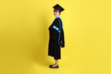 Mavi mezuniyet elbisesi ve şapkalı, ayakta durma ve kollarını çaprazlama hareketleriyle sarı arka planda izole edilmiş bir üniversite öğrencisi. Gen Z, yeni mezun, eğitim, ilk iş