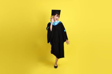 Sarı arka planda duran ve gülümseyen mavi mezuniyet elbisesi ve şapkalı bayan üniversite öğrencisi. Gen Z, yeni mezun, eğitim, ilk iş