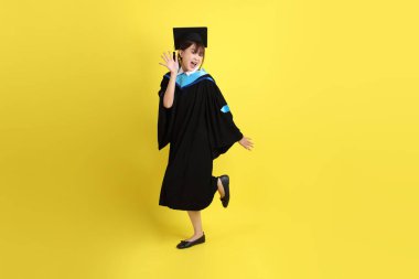 Sarı arka planda duran ve gülümseyen mavi mezuniyet elbisesi ve şapkalı bayan üniversite öğrencisi. Gen Z, yeni mezun, eğitim, ilk iş