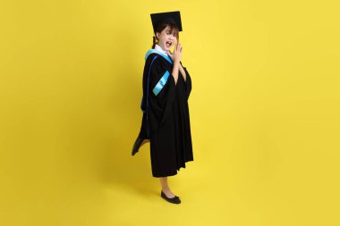 Sarı arka planda duran ve gülümseyen mavi mezuniyet elbisesi ve şapkalı bayan üniversite öğrencisi. Gen Z, yeni mezun, eğitim, ilk iş