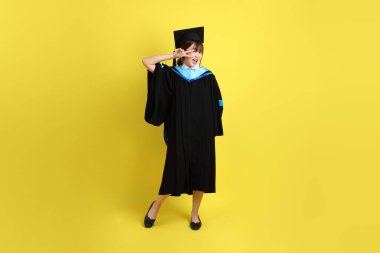 Mavi mezuniyet elbisesi ve şapkalı, barış işareti işareti olan bir üniversite öğrencisi. Sarı arka planda iki parmak izole edilmiş. Gen Z, yeni mezun, eğitim, ilk iş