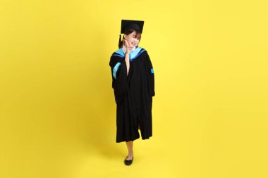 Mavi mezuniyet elbisesi ve şapkalı, barış işareti işareti olan bir üniversite öğrencisi. Sarı arka planda iki parmak izole edilmiş. Gen Z, yeni mezun, eğitim, ilk iş