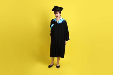 Mavi mezuniyet elbisesi ve şapkalı, sarı arka planda izole edilmiş düşünce jesti olan bir üniversite öğrencisi. Gen Z, yeni mezun, eğitim, ilk iş
