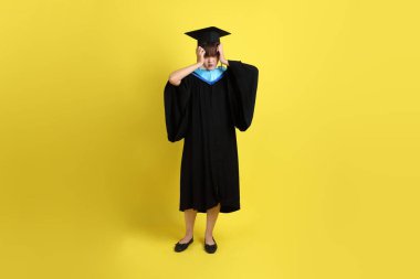 Mavi mezuniyet elbisesi ve şapkalı, sarı arka planda izole edilmiş düşünce jesti olan bir üniversite öğrencisi. Gen Z, yeni mezun, eğitim, ilk iş