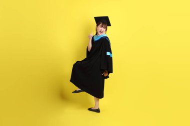 Mavi mezuniyet elbisesi ve şapkalı bayan üniversite öğrencisi, tamam ya da sarı arka planda izole edilmiş. Gen Z, yeni mezun, eğitim, ilk iş