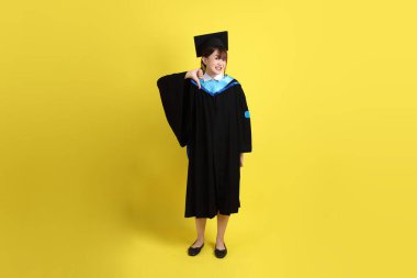 Mavi mezuniyet elbisesi ve şapkalı, sarı arka planda izole edilmiş baş parmağıyla hareket eden bir üniversite öğrencisi. Gen Z, yeni mezun, eğitim, ilk iş