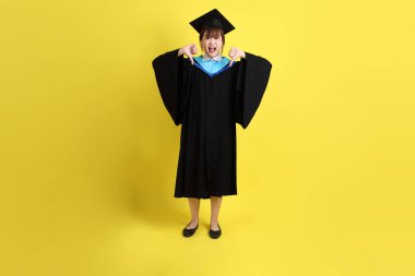 Mavi mezuniyet elbisesi ve şapkalı, sarı arka planda izole edilmiş baş parmağıyla hareket eden bir üniversite öğrencisi. Gen Z, yeni mezun, eğitim, ilk iş