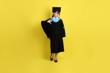 Mavi mezuniyet elbisesi ve şapkalı, sarı arka planda izole edilmiş baş parmağıyla hareket eden bir üniversite öğrencisi. Gen Z, yeni mezun, eğitim, ilk iş