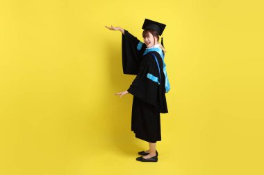 Mavi mezuniyet elbisesi ve kepleri içinde, sarı arka planda izole edilmiş bir şekilde işaret eden bayan üniversite öğrencisi. Gen Z, yeni mezun, eğitim, ilk iş