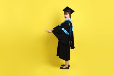 Mavi mezuniyet elbisesi ve kepleri içinde, sarı arka planda izole edilmiş bir şekilde işaret eden bayan üniversite öğrencisi. Gen Z, yeni mezun, eğitim, ilk iş