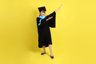 Mavi mezuniyet elbisesi ve kepleri içinde, sarı arka planda izole edilmiş bir şekilde işaret eden bayan üniversite öğrencisi. Gen Z, yeni mezun, eğitim, ilk iş