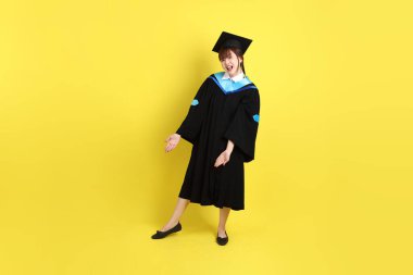 Mavi mezuniyet elbisesi ve kepleri içinde, sarı arka planda izole edilmiş bir şekilde işaret eden bayan üniversite öğrencisi. Gen Z, yeni mezun, eğitim, ilk iş