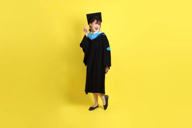 Mavi mezuniyet elbisesi ve kepleri içinde, sarı arka planda izole edilmiş bir şekilde işaret eden bayan üniversite öğrencisi. Gen Z, yeni mezun, eğitim, ilk iş