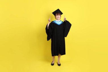 Mavi mezuniyet elbisesi ve kepleri içinde, sarı arka planda izole edilmiş bir şekilde işaret eden bayan üniversite öğrencisi. Gen Z, yeni mezun, eğitim, ilk iş