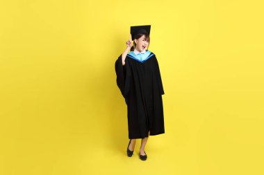 Mavi mezuniyet elbisesi ve kepleri içinde, sarı arka planda izole edilmiş bir şekilde işaret eden bayan üniversite öğrencisi. Gen Z, yeni mezun, eğitim, ilk iş