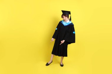 Mavi mezuniyet elbisesi ve kepleri içinde, sarı arka planda izole edilmiş bir şekilde işaret eden bayan üniversite öğrencisi. Gen Z, yeni mezun, eğitim, ilk iş