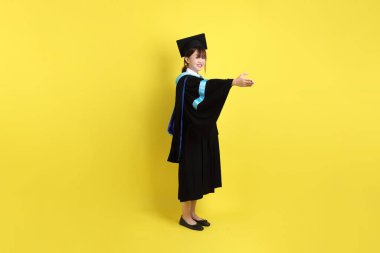 Mavi mezuniyet elbisesi ve kepleri içinde, sarı arka planda izole edilmiş, verme ve alma hareketi yapan bir üniversite öğrencisi. Gen Z, yeni mezun, eğitim, ilk iş