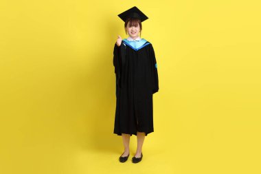 Mavi mezuniyet elbisesi ve kepleri içinde, sarı arka planda izole edilmiş, verme ve alma hareketi yapan bir üniversite öğrencisi. Gen Z, yeni mezun, eğitim, ilk iş