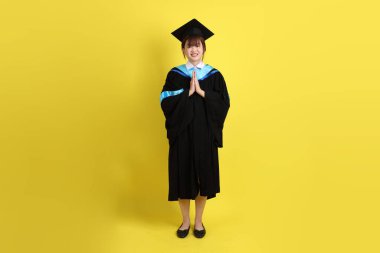 Sarı arka planda izole edilmiş selamlama jesti ile mavi mezuniyet elbisesi ve şapkalı bir üniversite öğrencisi. Gen Z, yeni mezun, eğitim, ilk iş