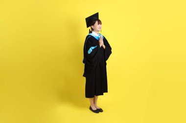 Sarı arka planda izole edilmiş selamlama jesti ile mavi mezuniyet elbisesi ve şapkalı bir üniversite öğrencisi. Gen Z, yeni mezun, eğitim, ilk iş