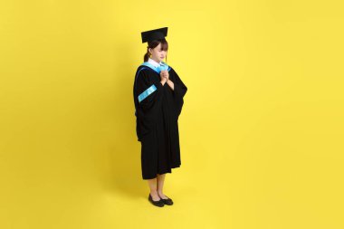 Mavi mezuniyet elbisesi ve şapkalı, sarı arka planda dua eden kız öğrenci. Gen Z, yeni mezun, eğitim, ilk iş