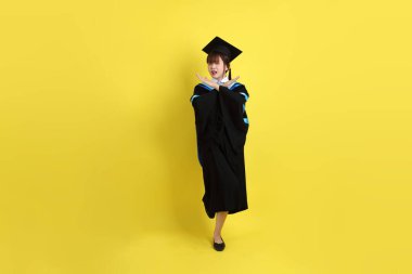 Mavi mezuniyet elbisesi ve şapkalı, sarı arka planda izole edilmiş, durdurulmuş, yasaklanmış ve reddedilmiş bir üniversite öğrencisi. Gen Z, yeni mezun, eğitim, ilk iş