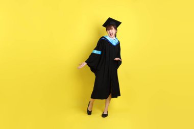 Mavi mezuniyet elbisesi ve kepleri içinde, sarı arka planda izole edilmiş bir şekilde işaret eden bayan üniversite öğrencisi. Gen Z, yeni mezun, eğitim, ilk iş