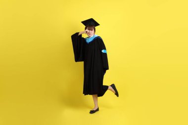 Mavi mezuniyet elbisesi ve kepleri içinde, sarı arka planda izole edilmiş, verme ve alma hareketi yapan bir üniversite öğrencisi. Gen Z, yeni mezun, eğitim, ilk iş