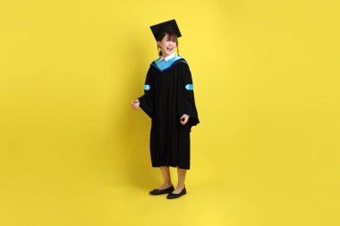 Sarı arka planda duran ve gülümseyen mavi mezuniyet elbisesi ve şapkalı bayan üniversite öğrencisi. Gen Z, yeni mezun, eğitim, ilk iş