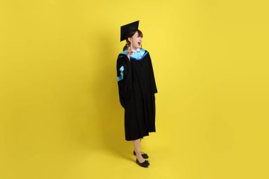 Mavi mezuniyet elbisesi ve kepleri içinde, sarı arka planda bir numaranın jesti olan bir üniversite öğrencisi. Gen Z, yeni mezun, eğitim, ilk iş