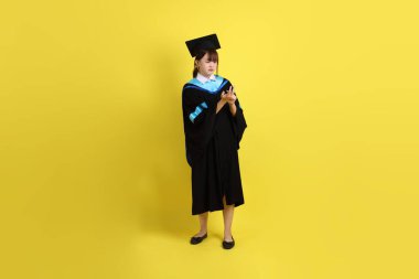 Mavi mezuniyet elbisesi ve kepleri içinde kadın üniversite öğrencisi. Sarı arka planda izole edilmiş sayım numarası ya da hesaplama jesti var. Gen Z, yeni mezun, eğitim, ilk iş