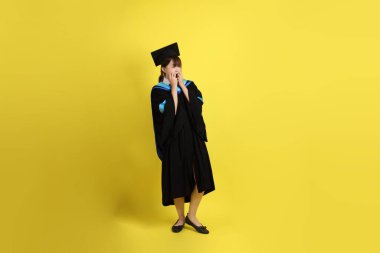 Mavi mezuniyet elbisesi ve şapkalı, sürpriz jestli ve sarı arka planda izole edilmiş etkileyici bir üniversite öğrencisi. Gen Z, yeni mezun, eğitim, ilk iş