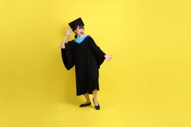 Mavi mezuniyet elbisesi ve şapkalı, sürpriz jestli ve sarı arka planda izole edilmiş etkileyici bir üniversite öğrencisi. Gen Z, yeni mezun, eğitim, ilk iş