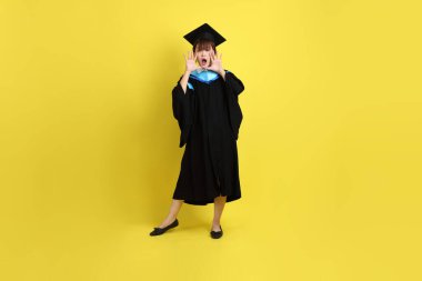 Mavi mezuniyet elbisesi ve şapkalı, sürpriz jestli ve sarı arka planda izole edilmiş etkileyici bir üniversite öğrencisi. Gen Z, yeni mezun, eğitim, ilk iş