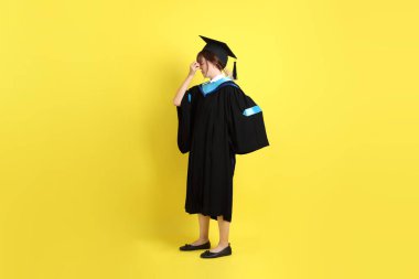 Sarı arka planda izole edilmiş stres jesti ile mavi mezuniyet elbisesi ve kepleri içinde bayan üniversite öğrencisi. Gen Z, yeni mezun, eğitim, ilk iş